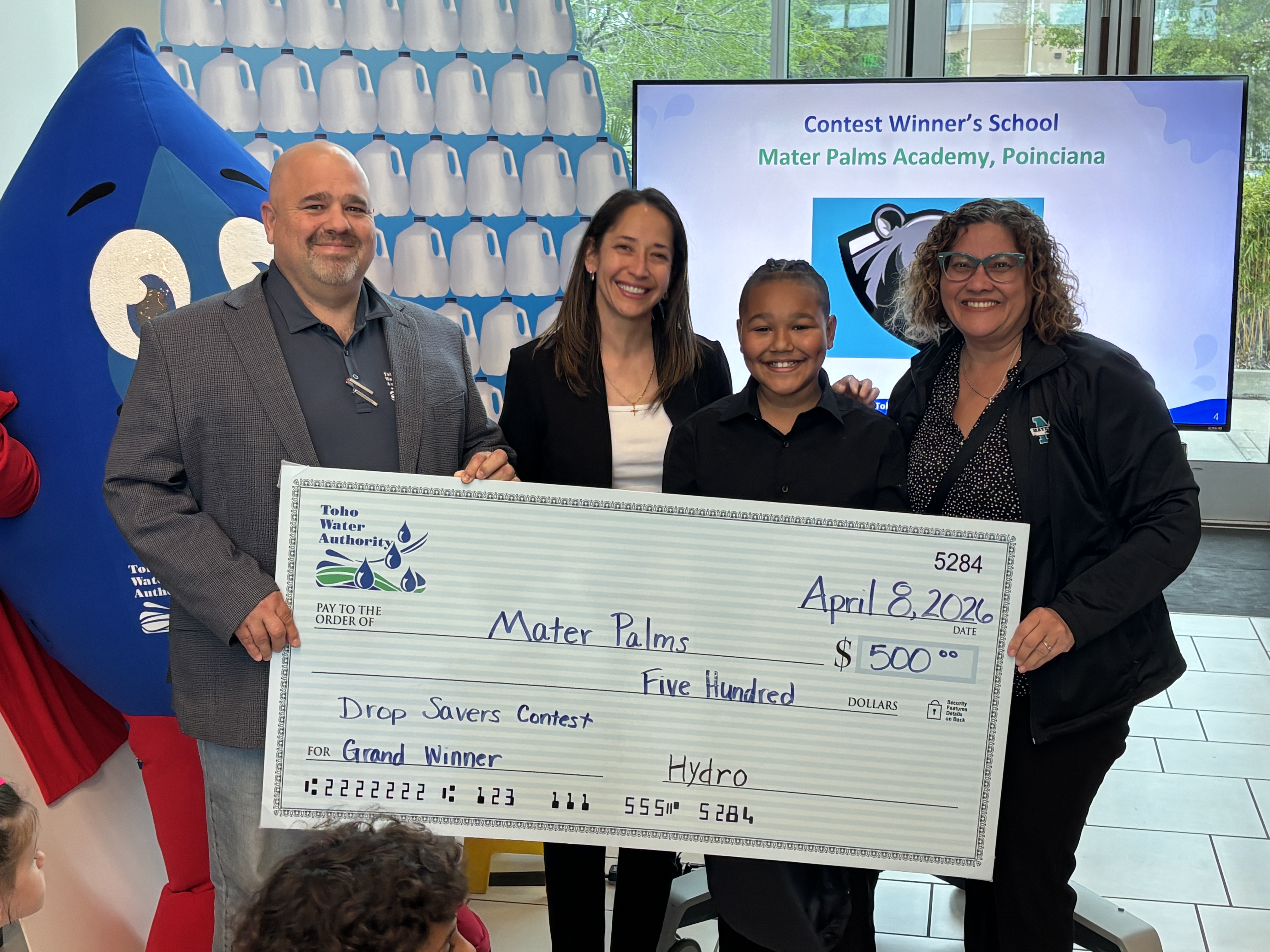ceo presents large check to a student and their teacher with a blue water drop mascot in front of a television saying contest winner's school, mater palms academy poinciana