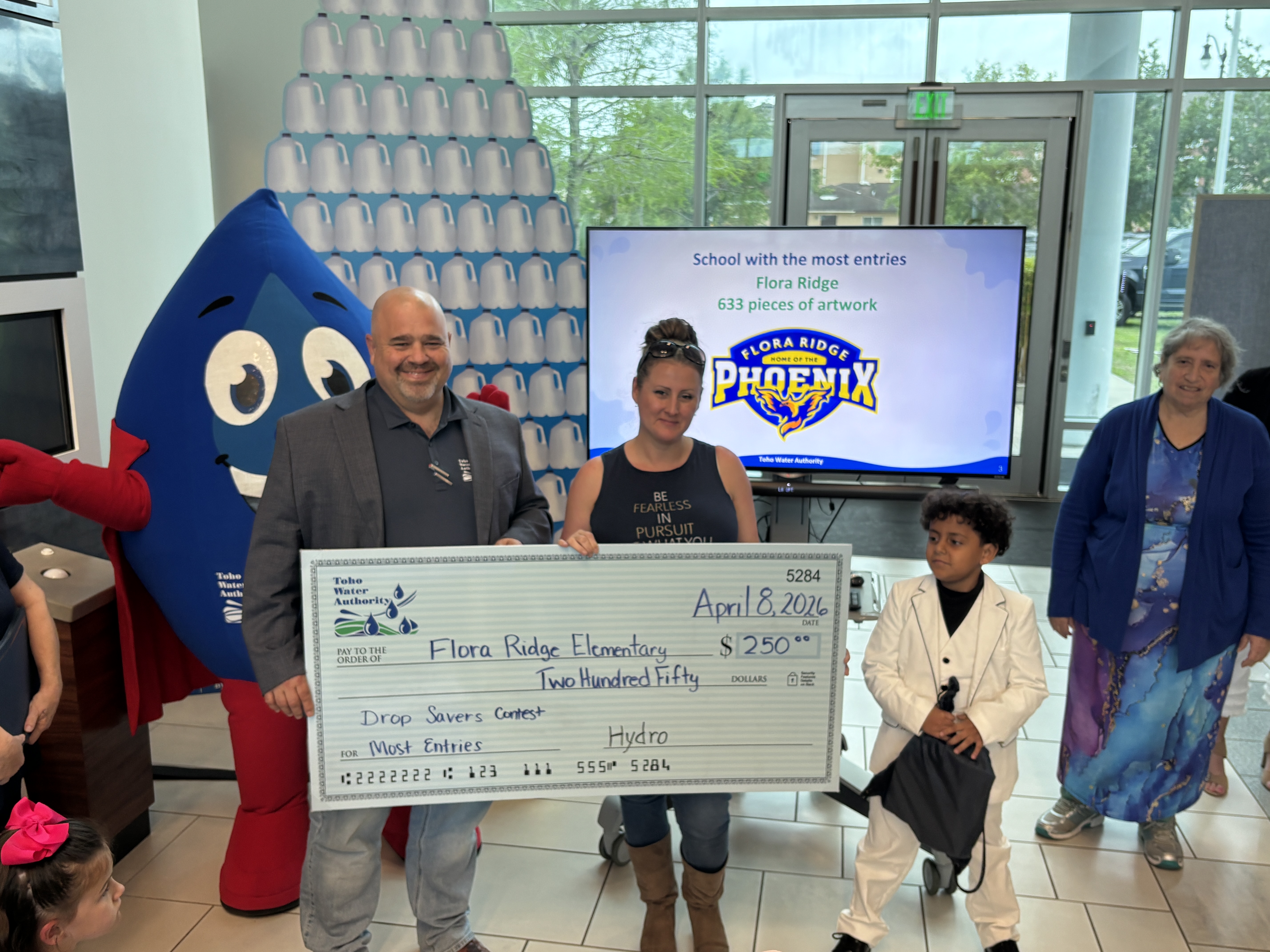 blue water drop mascot poses with ceo who presents a check to an adult female teacher with a young boy students and an adult woman in front of a screen that says school with the most entries flora ridge 633 pieces of artwork with a school logo