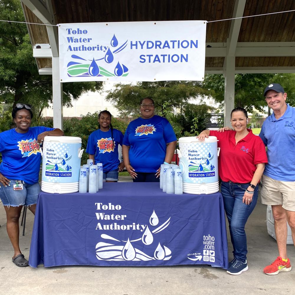 Photo of five Toho team members standing next to a table with two water coolers and cups. A banner with the Toho logo is hanging with the words "Hydration Station."