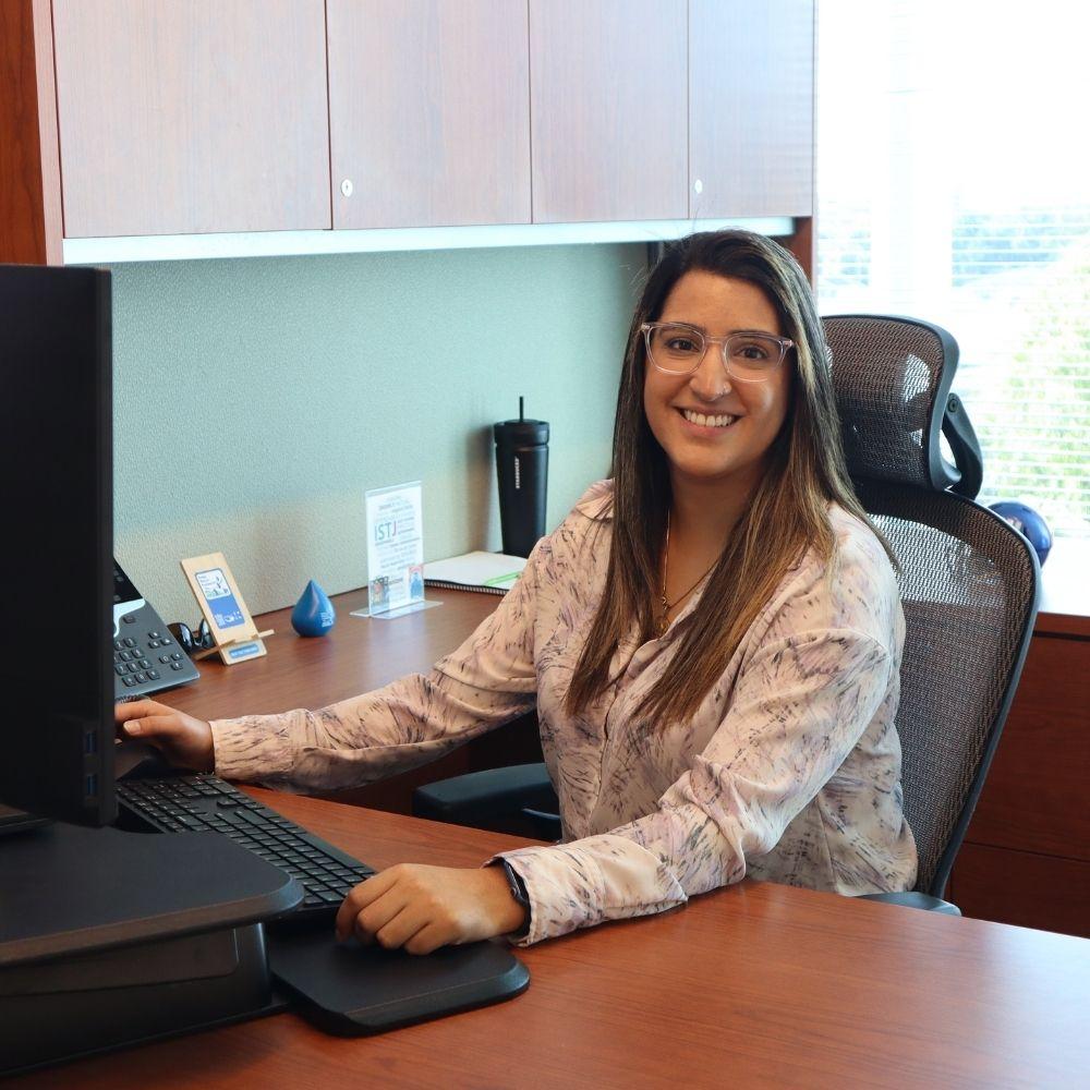 Image of female employee sitting at her desk computer in an office smiling at the camera.  
